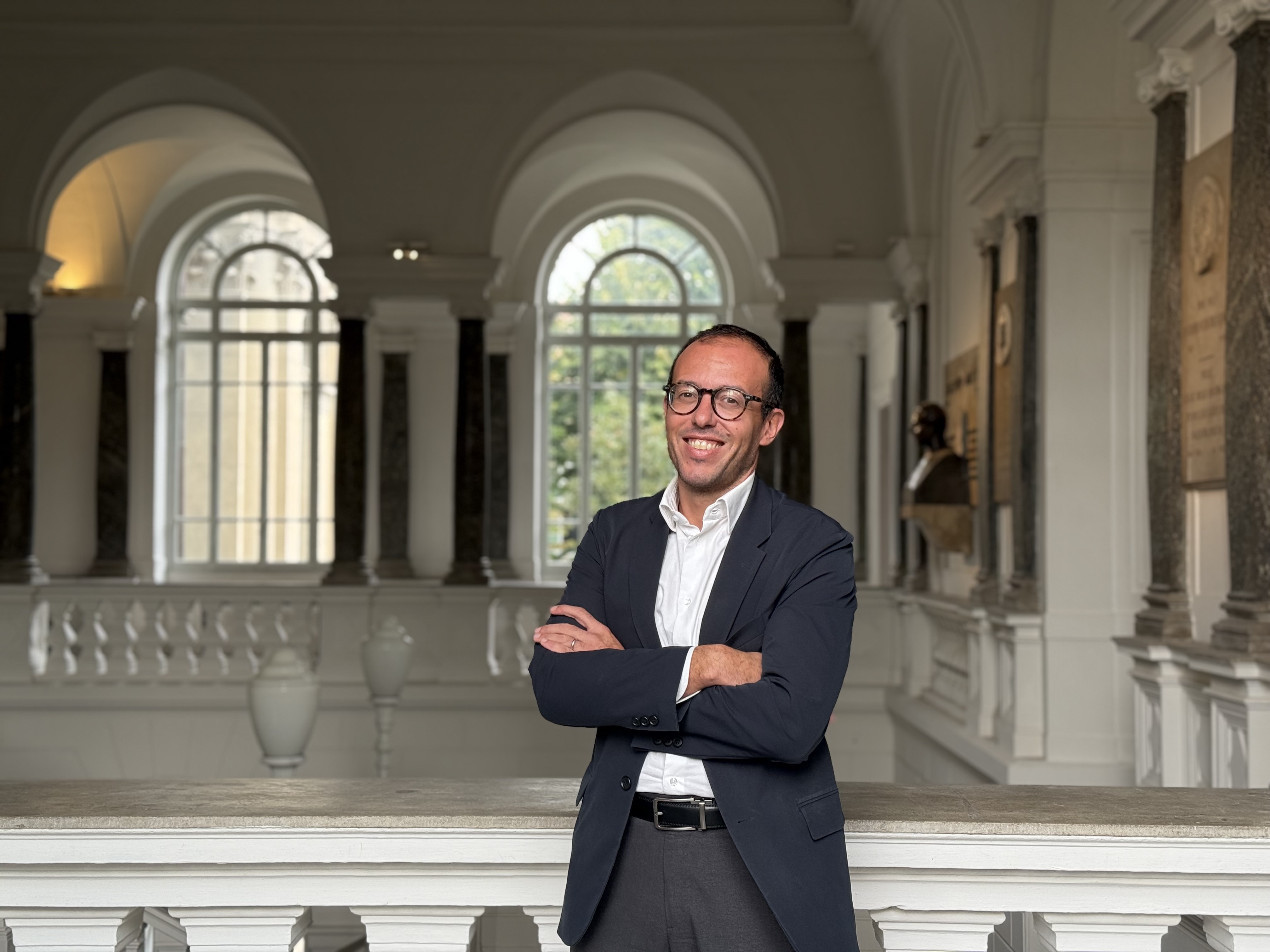 Manager sulla quarantina, capelli neri, occhi marroni e occhiali rotondi. Sorride e ha le braccia incrociate. indossa una camicia bianca e una giacca da vestito blu scuro. Si trova in una balconata interna, con due finestre ad arco che fanno da sfondo.